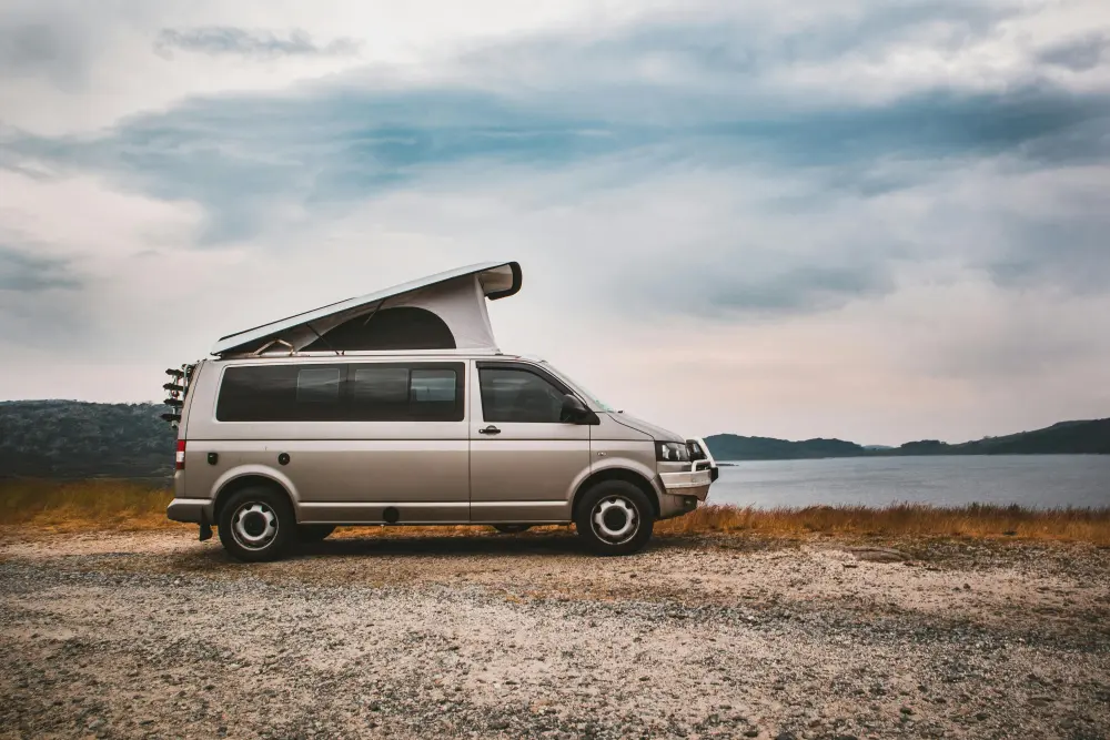 Ein Ford Transit Custom Campervan mit hochgeklapptem Dach steht an einem Seeufer unter bewölktem Himmel, perfekt für Outdoor-Abenteuer und Camping.